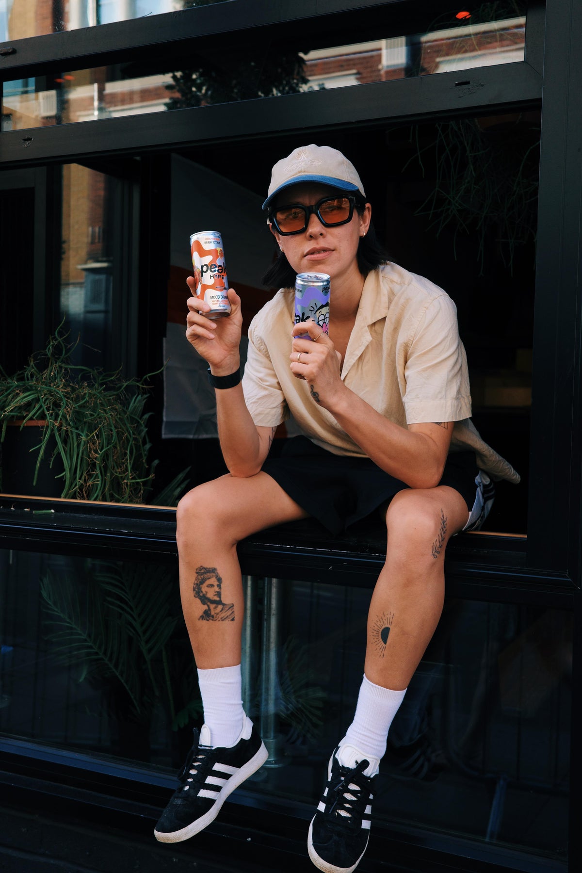 A person in a cap and sunglasses sitting on a window ledge holding a can of Peak Hype, a drink with 250mg of L-tyrosine, and a can of Peak Calm